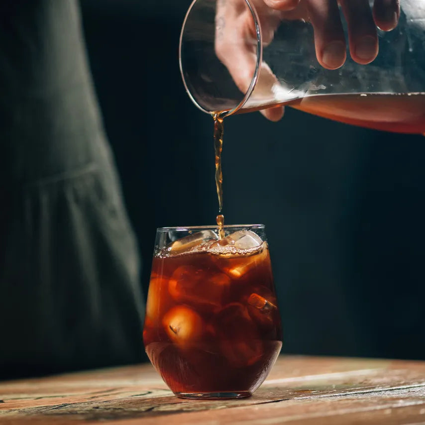 Person pouring a clear container of cold brew into a glass with ice on a wooden surface.