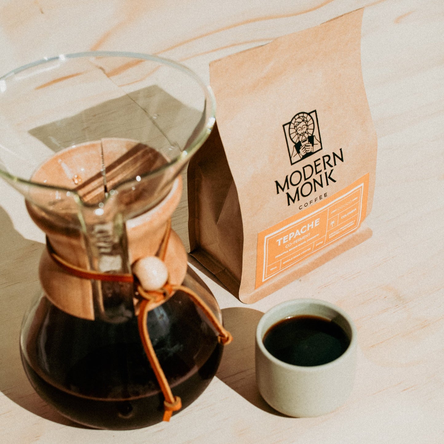 Chemex coffee maker with a cup of coffee and a bag of Modern Monk Coffee on a wooden surface.