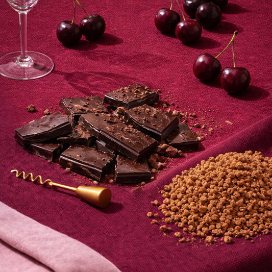 Dark chocolate pieces, cherries, and a glass of red wine on a red surface.