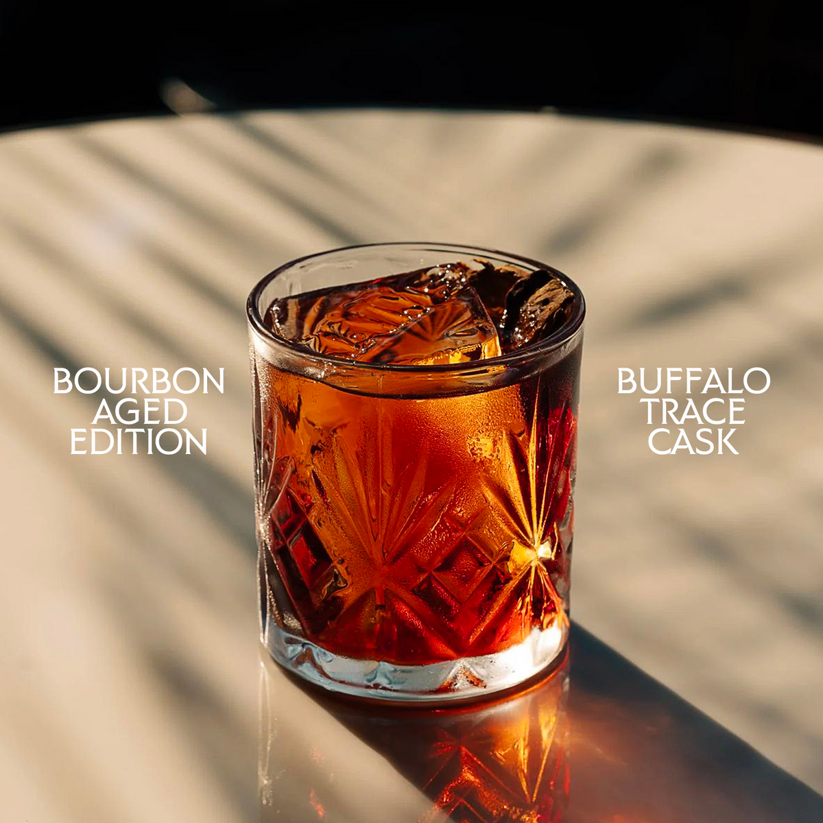A clear whiskey tumbler with a starburst cut pattern, filled with an amber-brown bourbon cocktail and ice, set on a reflective surface with dramatic shadows. White text on the left reads 'BOURBON AGED EDITION' and text on the right reads 'BUFFALO TRACE CASK'.