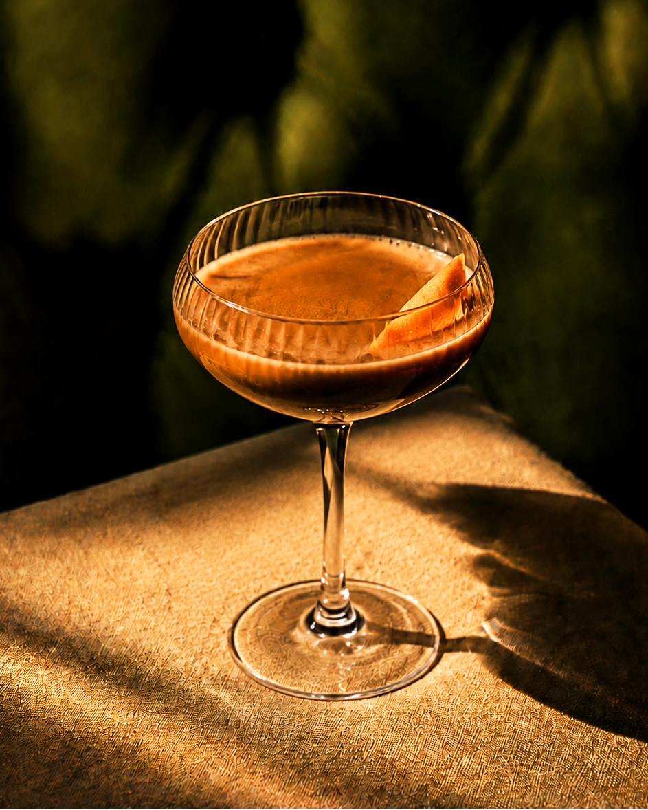 A coupe glass filled with an orange-brown cocktail, garnished with a citrus peel, set on a textured surface under dramatic warm lighting against a dark green background.