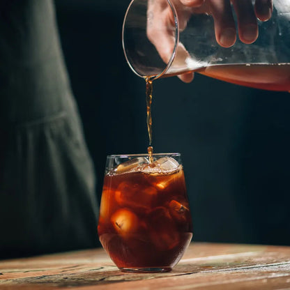 Person pouring a clear container of cold brew into a glass with ice on a wooden surface.
