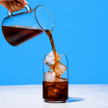 Hand pouring iced coffee from a pitcher into a glass against a blue background