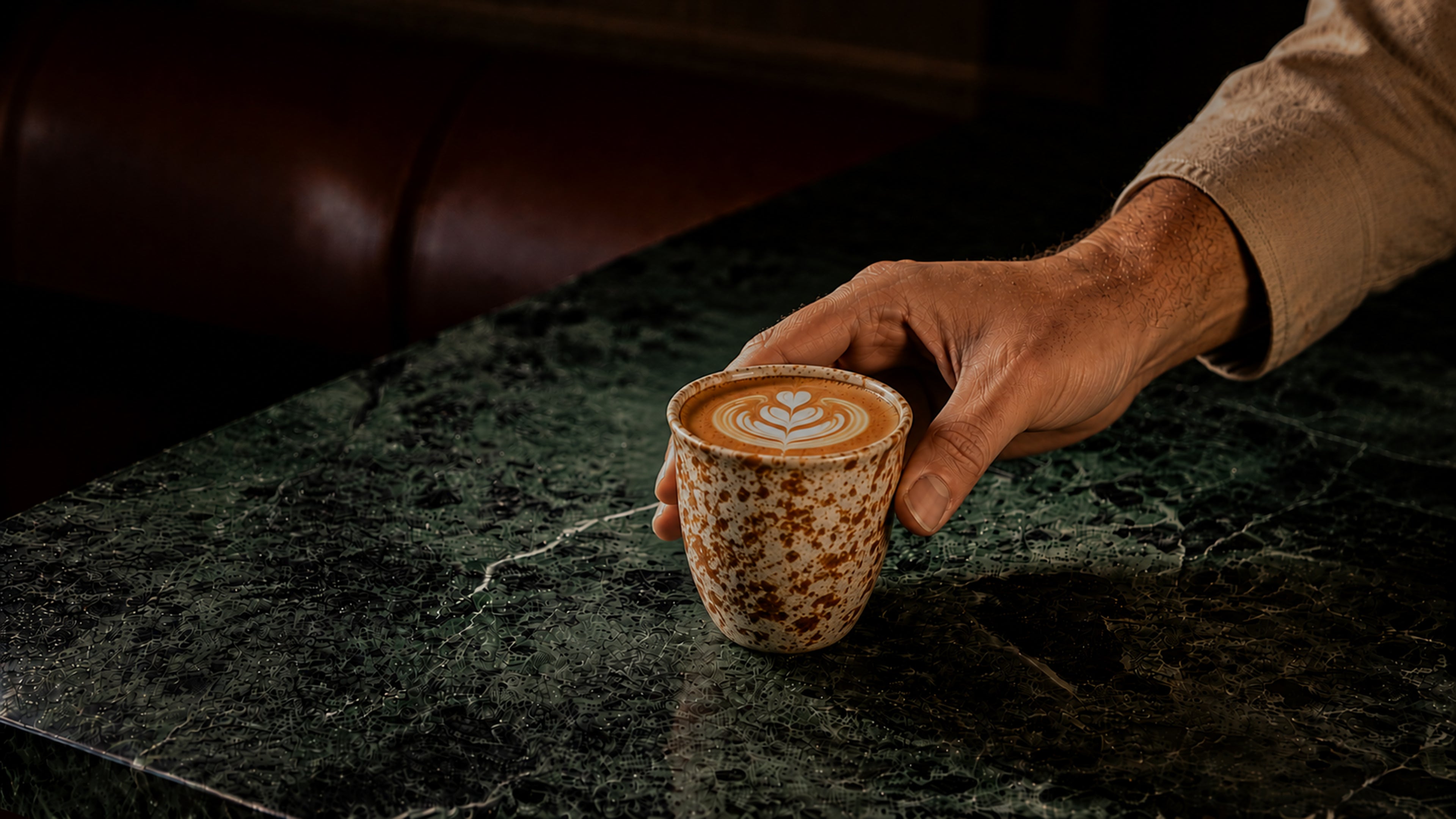Hand holding a small ceramic cup with a latte art design on a dark surface.